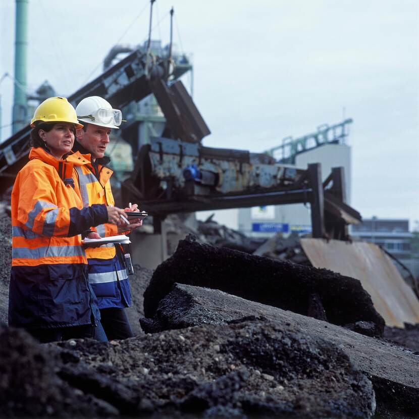 Twee mensen in oranje veiligheidsjassen en helmen staan op een bouw- of recyclinglocatie. Op de voorgrond liggen grote stukken asfalt of bouwpuin, terwijl op de achtergrond een machine zichtbaar is die materiaal verwerkt.