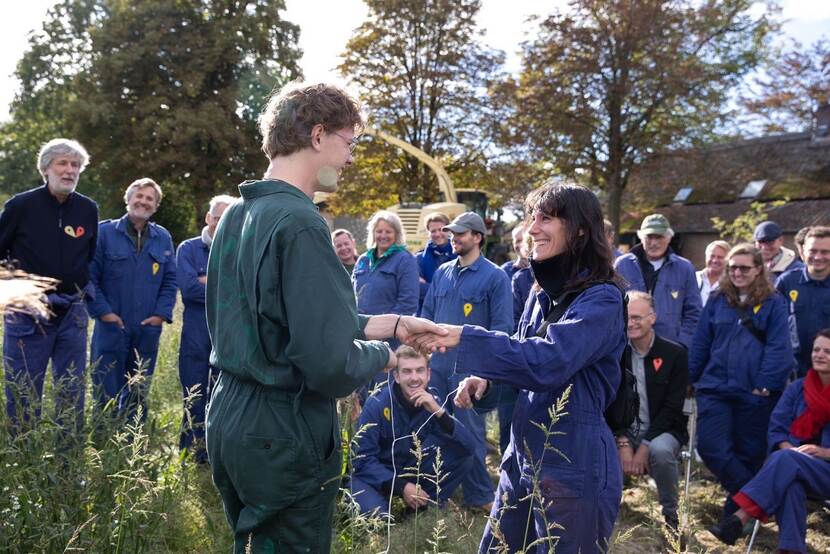 Een groep mensen in blauwe en groene overalls staat buiten op een grasveld en kijkt glimlachend toe terwijl twee mensen elkaar de hand schudden.