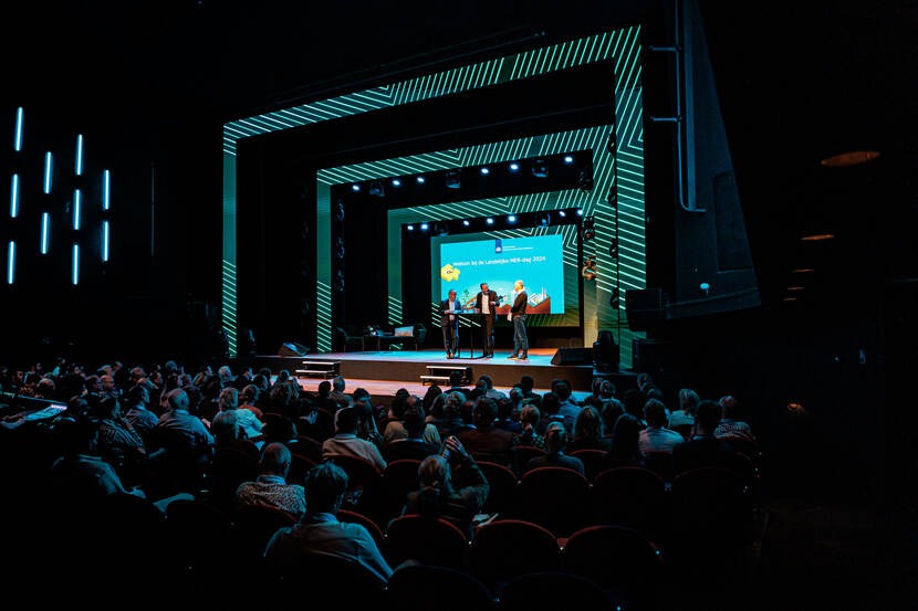 Een volle theaterzaal tijdens de Landelijke MER-dag 2024. Op het podium staan drie sprekers voor een groot scherm met de tekst "Welkom bij de Landelijke MER-dag 2024."